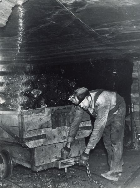 A miner fastening a chain to a mining car in order to be pulled from the shaft by a drum hoist. The drum hoist was powered by an International U-7 power unit.