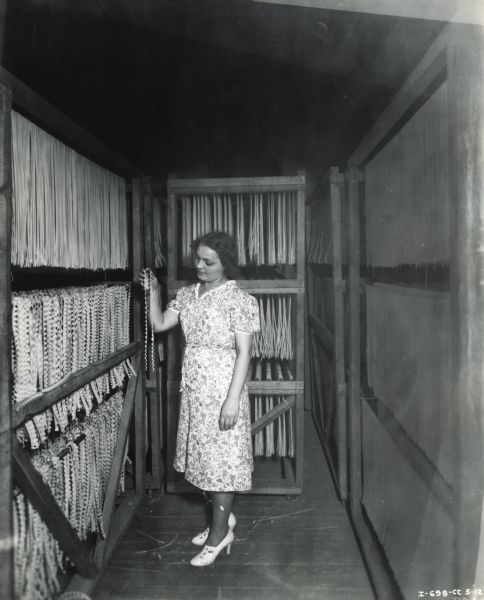 A woman wearing a dress and high-heeled shoes holds a strand of macaroni in a room at the Florida Macaroni Company. Various types of macaroni products hang on wooden drying racks around her. An International P-12 power unit was used to operate all machinery in the factory.
