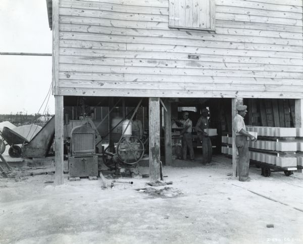 Men loading cement blocks onto rolling carts in an open-air building at Naranja Rock Company Incorporated. An International Model 200 power unit was used to power various machinery.