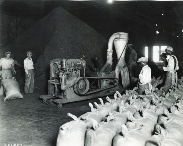 Workers gather around an International Harvester Model 1-B hammer mill powered by an International Model 200 power unit at the Fish Uleal Company.  The machines were used to grind fish for fertilizer; burlap bags are stacked in the foreground.