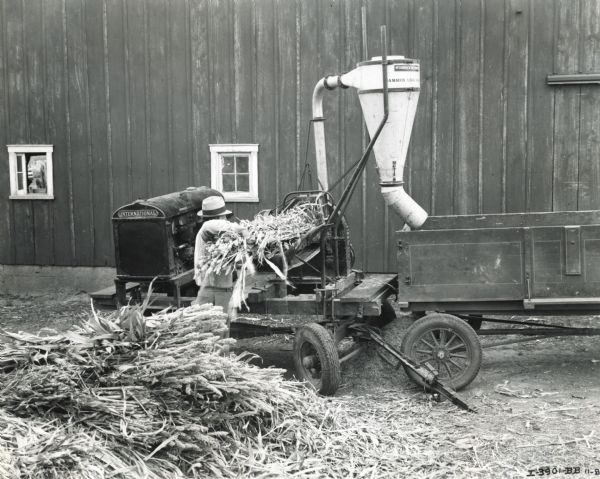 A man loads what appear to be stalks of harvested wheat onto a #10-C hammer mill operated by a P-30 power unit. The entire outfit was mounted on a tractor-trailer and towed by a D-2 International truck.