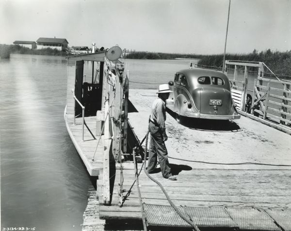 A man stands holding a rope on an auto ferry dock. The ferry was powered by an International P-12 power unit and provided transportation to Ridge Island. An automobile is parked on the ferry and buildings are across a body of water in the background.
