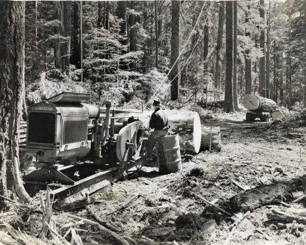 A man sits on a metal barrel near an International I-30 tractor modified into a P-30 power unit. The power unit was used with a Skagil hoist at a lumber site in a forested area. The power unit was owned by Charles Holmes.