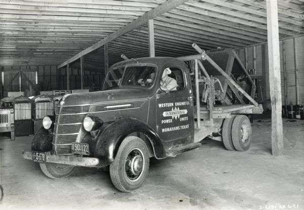 A man sits behind the wheel of an International D-15 truck owned by Western Engine Company and used to make deliveries of power units.  The photograph is taken inside a storage building with power units in the background.