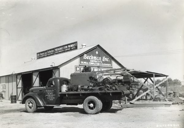 International D-50 truck with an International power unit loaded on the bed. The truck is parked in a dirt lot in front of Beckman Incorporated Contractors and Muskogee Iron Works building.