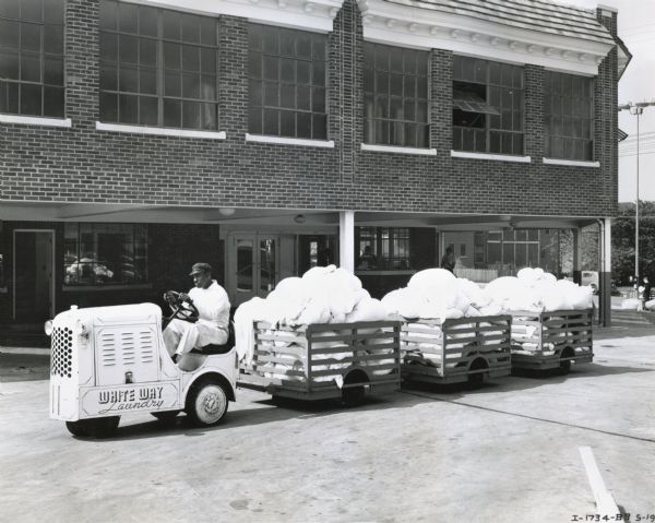 A man uses a Hebard shop mule owned by White Way Laundry to haul three-wheeled bins of bagged laundry. The shop mule, powered by an International P-12 power unit, is moving through a parking lot in front of what appears to be the laundromat building.