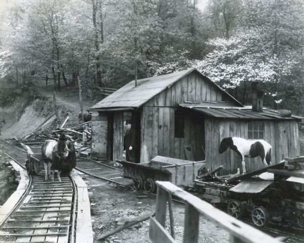 Men and ponies standing near a building and carts on railroad tracks at the Story & McDade coal mine. The mine operation was powered by an International PA-40 power unit and a 30-horsepower generator.