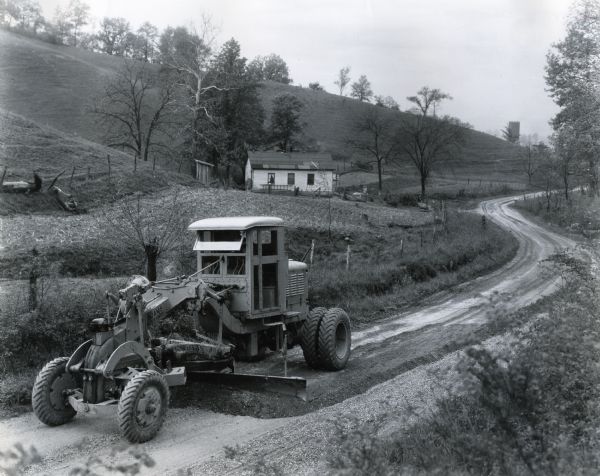 An Adams road grader equipped with a McCormick-Deering power unit is used by a county road crew to smooth a road in a rural area near the Ohio and Indiana border. A building is in the background.