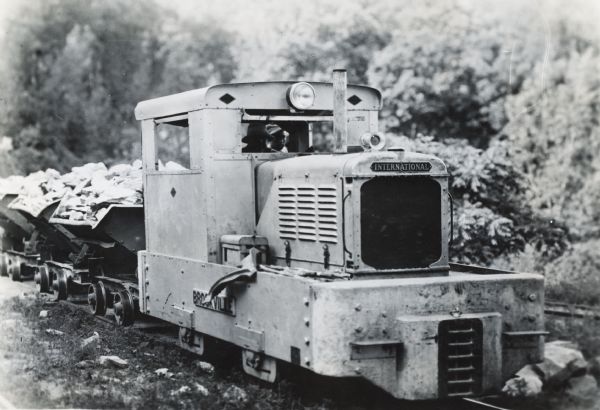 Brookville locomotive pulling dump cars in a rock quarry owned by General Refractories Company. Two men can be seen sitting inside through the front window of the locomotive. The locomotive was powered by an International PD-40 power unit.