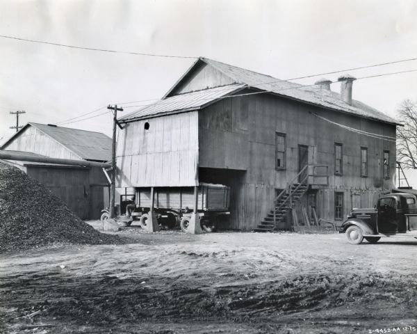 Exterior view of the Rosedale Gin, powered by an International PD-80.  Two trucks are parked near the building. One of the trucks has a wagon attached.