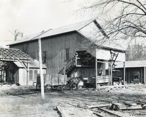 Exterior view of a cotton gin owned by O.L. Gormon. A man stands on a balcony under the overhang of the building at the top of a flight of steps. Another man stands in the back of a wagon pulled by a truck, which is parked under the same overhang; another horse-drawn wagon waits behind.
