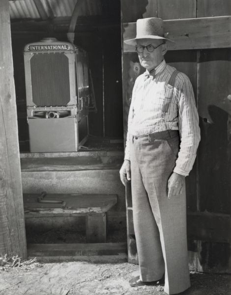 Mr. F.M. Smith, in a bow tie, hat, and trousers with suspenders, stands near the open door of a barn or shed. Inside is his International P-12 power unit.