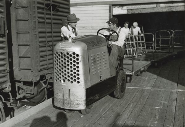 H.H. Harrell, seated behind the wheel of an International-powered shop mule, receives instructions from the general foreman, H.J. McClusky.  The text on the shop mule reads: "50 PAC Lines; Los Angeles." In the background in a building and two other men are standing near the carts that are being hauled by the shop mule. There is a railroad car on the left.
