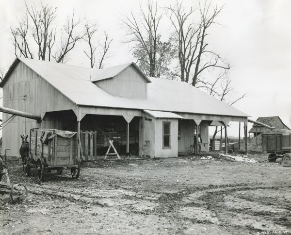 Exterior view of a cotton gin owned by R.E. Cox. Two men stand beneath the building's overhang and a mule or horse-drawn wagon stands to the left of the building.