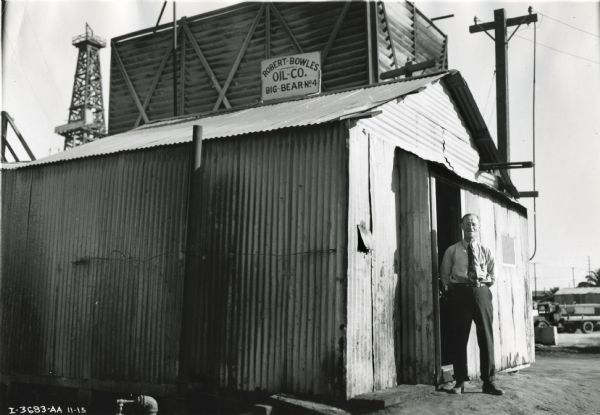 A man stands near the opened door of the corrugated shed of the Robert Bowles Oil Company. The company used an International Model 300 power unit to power the oil well.