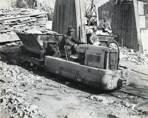 A man operating a Brookville locomotive powered by an International P-12 power unit while two men are working behind him at what appears to be a construction site.