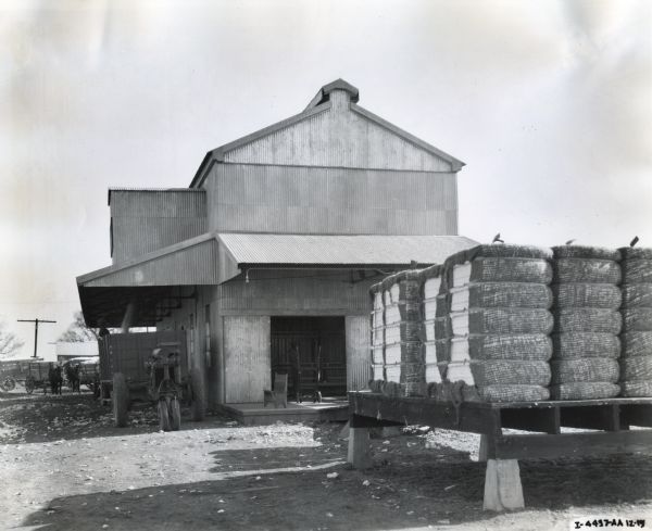 Exterior view of the cotton gin at Siut-Me Gin Company. Bales of cotton are piled on a platform in the foreground, and horse-drawn and tractor-drawn wagons are waiting in line on the left to unload under the covered roof of the building.
