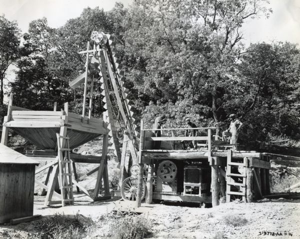 Three men stand on a platform to operate a rock crusher powered by an International PA-50 power unit, which is underneath the platform. The photograph was taken in Adair County.