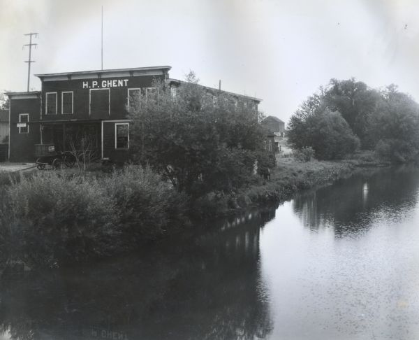 Elevated exterior view of the H.P. Ghent woodworking shop. A horse stands in the grass near a shoreline on the right, and a wagon parked near the shop bears a sign reading: "Samara Coal."