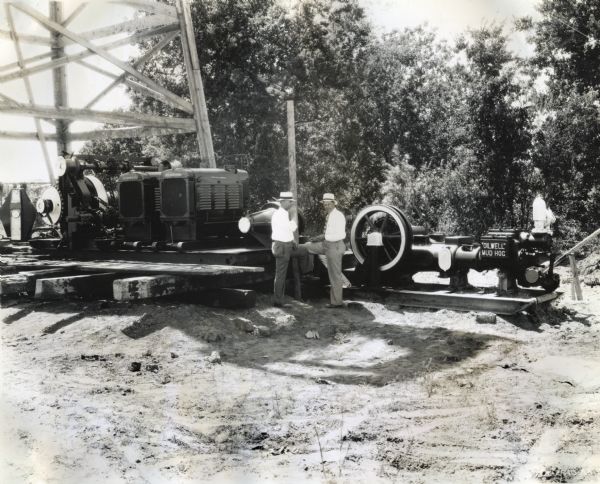 Two men wearing white hats stand near a drilling rig on which two International PA-50 power units are compounded. The machine on the right has a sign reading: "'Oil Well' Mud Hog."
