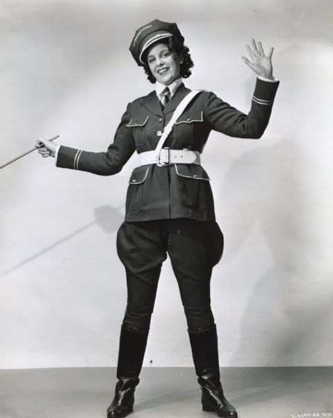 A woman stands in front of a white backdrop holding a baton. She is wearing a traffic officer costume with a hat and boots for an International Harvester Company road show.