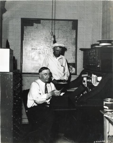 George J. Landaal sits at a large wooden desk and examines a piece of paper taken from a box in the Landaal Brothers office while Alfred Faller looks on over his shoulder. A map hangs on the wall in the background. Landaal Brothers was an International Harvester dealership.