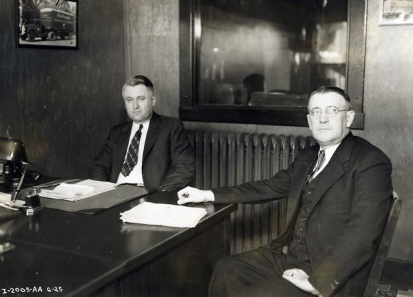E. Watson (left), manager of freight operations, discusses plans for expansion with Walter Hitchin, general freight agent, in an office. A photograph of a truck hangs on the wall to the left of the men. The office may be part of an International dealership or branch.