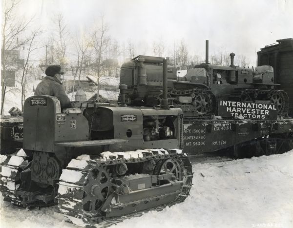 A man operates an International TD-35 TracTracTor (crawler tractor) on snow-covered ground as additional TracTracTors are unloaded from a train car marked with a sign reading: "International Harvester Tractors." The photograph was taken in Hudson, Ontario, Canada.