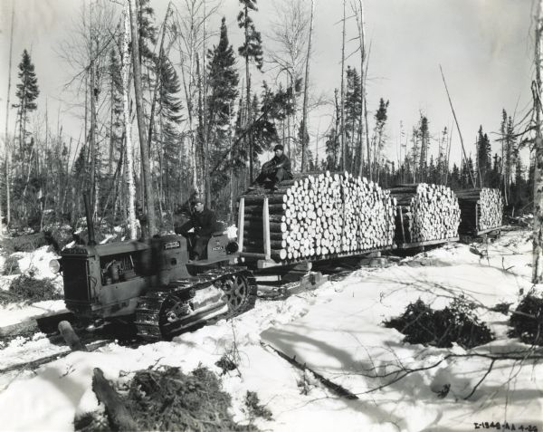 Two men use an International Model TD-35 TracTracTor (crawler tractor) to haul sleds piled with logs across snow-covered ground in a wooded area. The photograph was taken in Fort Frances, Ontario, Canada.