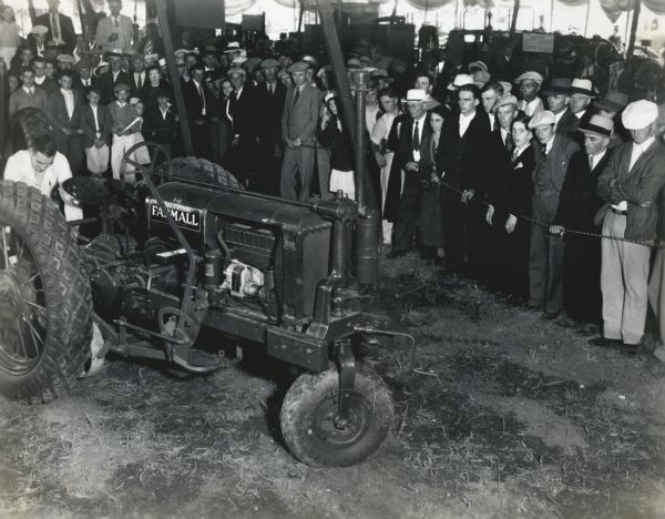 A crowd of onlookers gathers underneath a tent to watch a demonstration of a Farmall F-12 tractor with a single front wheel at the Wisconsin State Fair.  Other farm equipment is in the background.