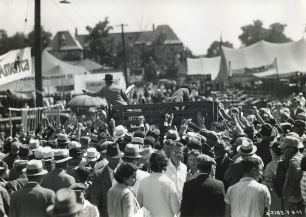 A crowd gathered around two men standing in an open wagon at the Indiana State Fair. Many of the people in the crowd have their hands raised. A sign reading "America" and tents are in the background. The men are likely handing out souvenir yardsticks on behalf of the International Harvester Company.