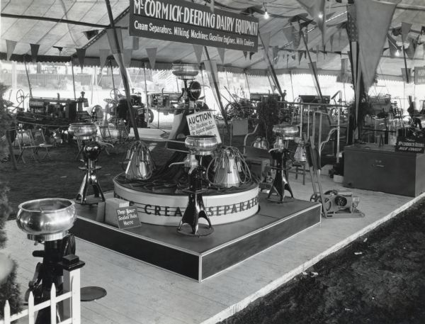 A cream separator is up for auction as part of a display of McCormick-Deering dairy equipment at the Indiana State Fair. Additional farm equipment is in the background and decorative pennants and flags hang from the tent ceiling. The sign hanging from the ceiling reads: "McCormick-Deering Dairy Equipment; Cream Separators, Milking Machines, Gasoline Engines, Milk Coolers." The signs on either side of the cream display read: "Drop Your Sealed Bids Here" and "Auction: This machine to the highest bidder."