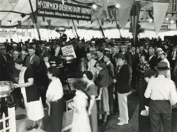 A crowd of people gathers in the McCormick-Deering dairy equipment display tent at the Indiana State Fair. The sign hanging from the ceiling reads: "McCormick-Deering Dairy Equipment; Cream Separators, Milking Machines, Gasoline Engines, Milk Coolers."