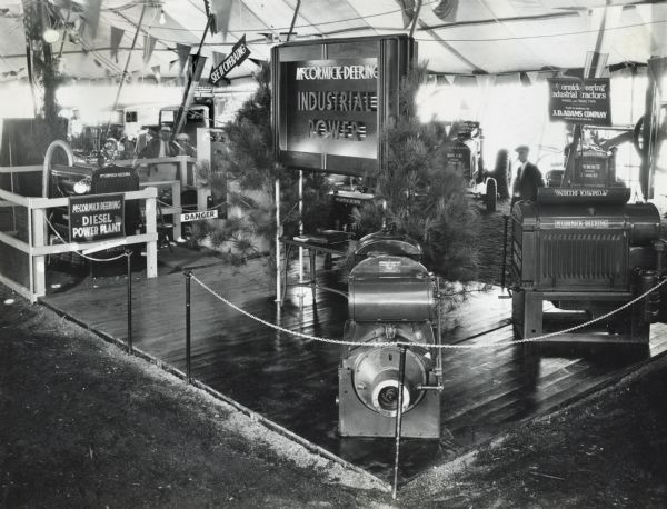 Engines and power units on display beneath a tent at the Indiana State Fair. An electric sign flanked by two evergreen trees reads: McCormick-Deering Industrial Power."