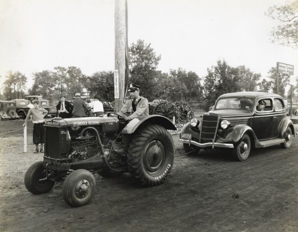 A uniformed man, possibly a police officer or a security guard, drives a McCormick-Deering I-12 tractor through the Eastern States Exposition grounds towing an automobile in which a similarly dressed man sits in the driver's seat. A boy and several men stand watching behind the vehicles.