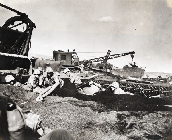 Marines dig foxholes into the sand on the beaches of Iwo Jima. The men are surrounded by military equipment, including a crane. An International Diesel crawler tractor (TracTracTor) is in the background.