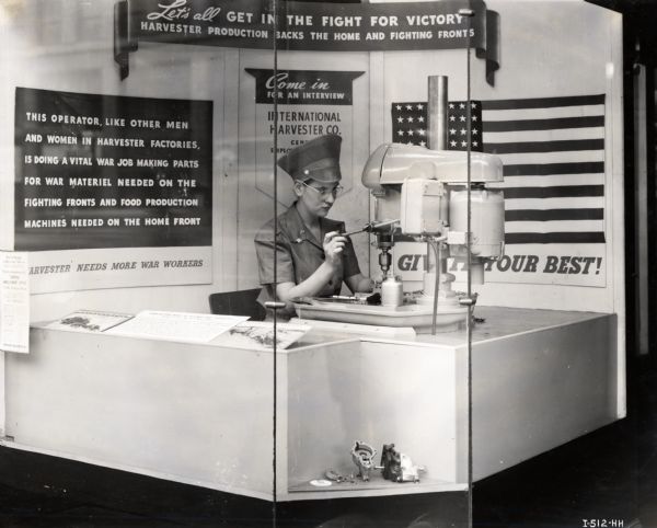 A woman sits behind a glass wall while operating a piece of machinery as part of an International Harvester display recruiting additional workers during World War II.
