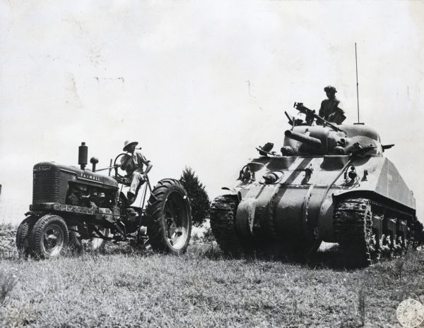 A man wearing a hat and smoking a pipe sits on a Farmall H tractor while a uniformed man stands on the top of a military tank. Two more men are sitting in the front of the tank.
