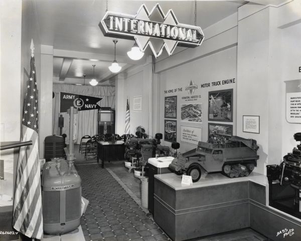Interior view of the Labor Recruiting Office of International Harvester's Indianapolis Works (factory). The office features models of military vehicles, American flags, a neon sign hanging from the ceiling, and a wall display of photographs. In the back of the room is a banner that reads "ARMY E NAVY" in front of rows of folding chairs and a television.