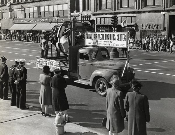 Men and women stand on either side of a street to watch an International truck used as a float for the Naval Air Tech Training Center as part of a Navy Day parade. The signs on the side of the float read, "Quick Engine Change Stand" and "Naval Air. Tech. Training Center; 87th and Anthony; Where the Navy Trains the Men Behind the Men With Wings."