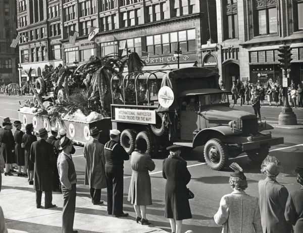 Me and women line the sidewalks to watch an International Harvester parade float go by. The float is decorated with uniformed men in a jeep, and palm trees; the sign on the float reads, "International Harvester Builds Trucks For the Navy."