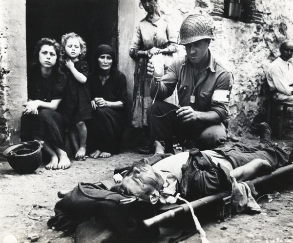 Pfc. Harvey White of Minneapolis, Minnesota gives blood plasma to a wounded soldier lying on a stretcher, while civilian women and children look on from the doorway of a nearby building. The photograph was taken in Sicily, Italy.