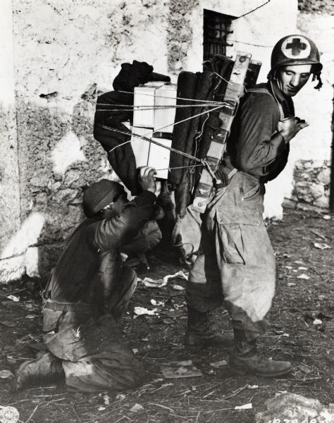 A man kneels to secure the ties on equipment strapped to the back of a medic wearing a Red Cross helmet.