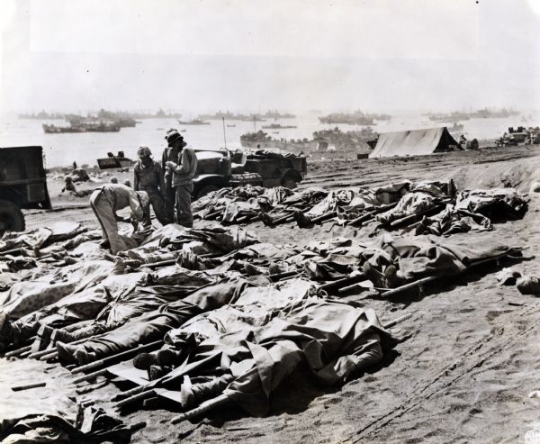 Marines, killed in action, lie on stretchers covered with ponchos on the beach at Iwo Jima. Four marines attend to the dead. Tents, military boats, and an International truck are in the background.