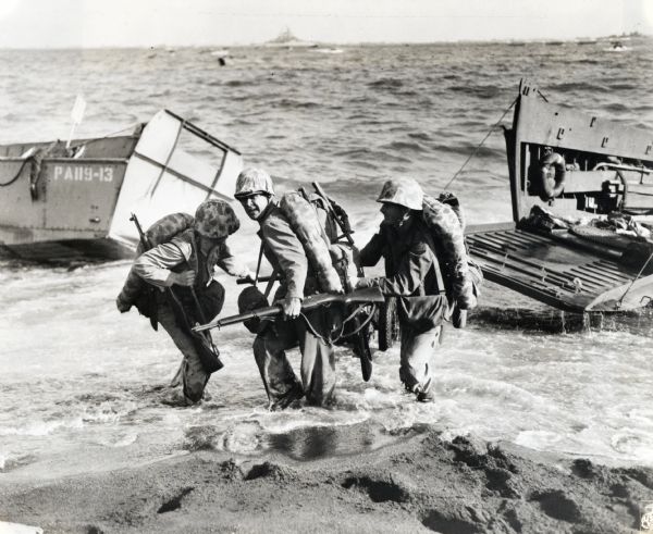Three marines dressed in uniform, carrying packs and holding guns, pull an ammunition cart through the water towards the beach at Iwo Jima. Military boats are in the background.