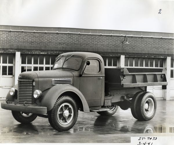 View of the driver's side of an International K-7 4x2 truck with a dump body made for the U.S. Army-Quartermaster Division. The truck is parked on a paved lot in front of what appears to be a garage.