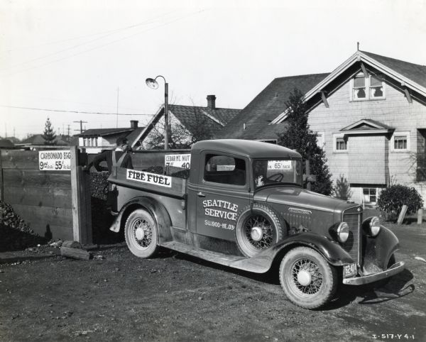 International C-1 truck with a pickup body owned by the Fire Fuel Company is parked while a man shovels cargo from a wooden bin onto the truck bed. There are signs on each bin of coal describing type and price. The Text on the truck reads: "Fire Fuel" and "Seattle Service." Houses are in the background.