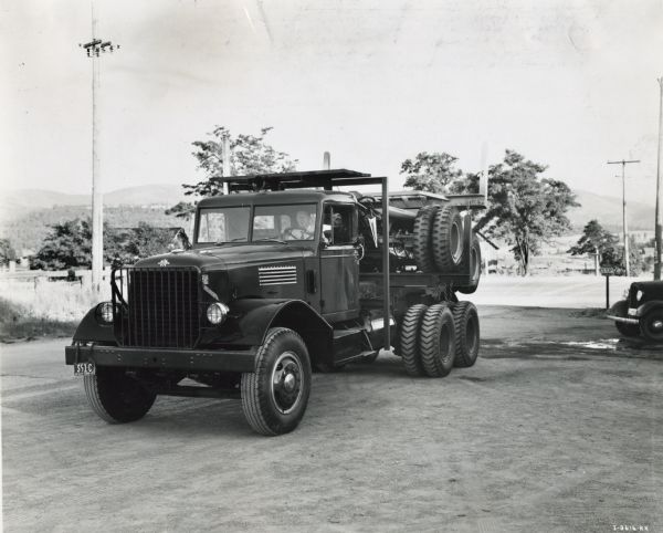 Three-quarter front view of the driver's side of an International truck parked on a dirt surface with a man at the wheel. The bed of the truck is loaded with what appears to be a long log trailer.