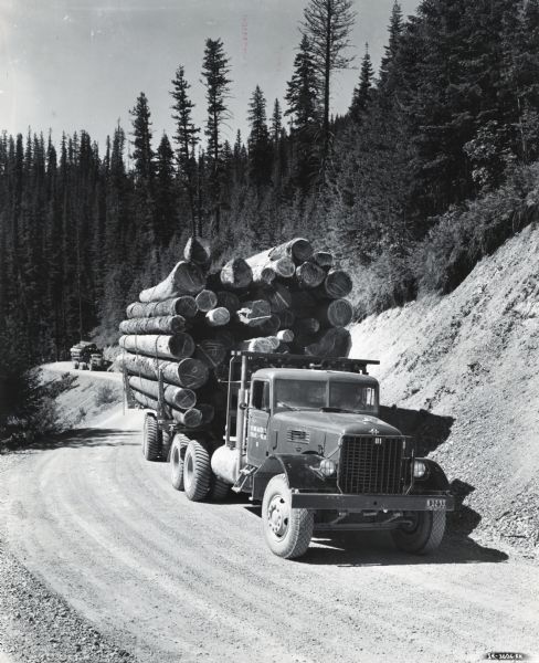 An International truck owned by the Ohio Match Company carries a load of logs down a gravel road in a wooded area in the vicinity of Coeur d'Alene. Another truck carrying logs is in the background.