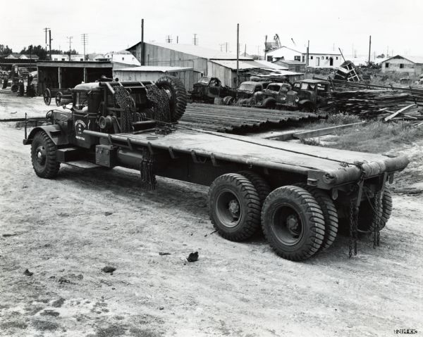 Slightly elevated view of an International truck with a flatbed and winch(?) owned by Joe Hughes Incorporated. The truck is parked in a dirt lot and storage buildings, refuse and scrapped automobiles are in the background.
